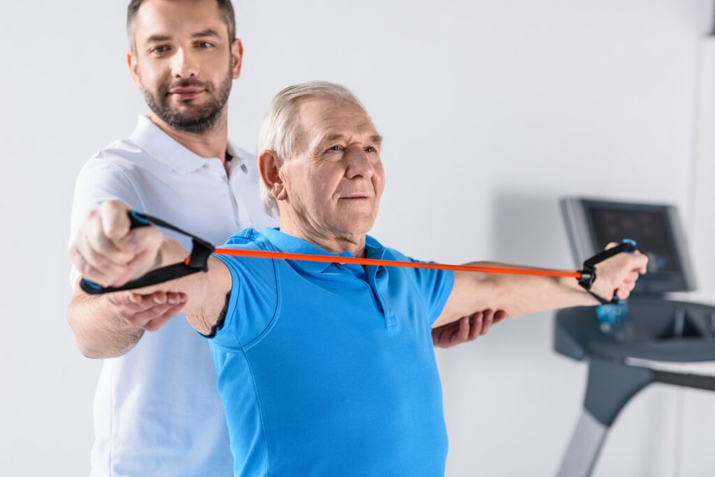 smiling rehabilitation therapist assisting senior man exercising with rubber tape